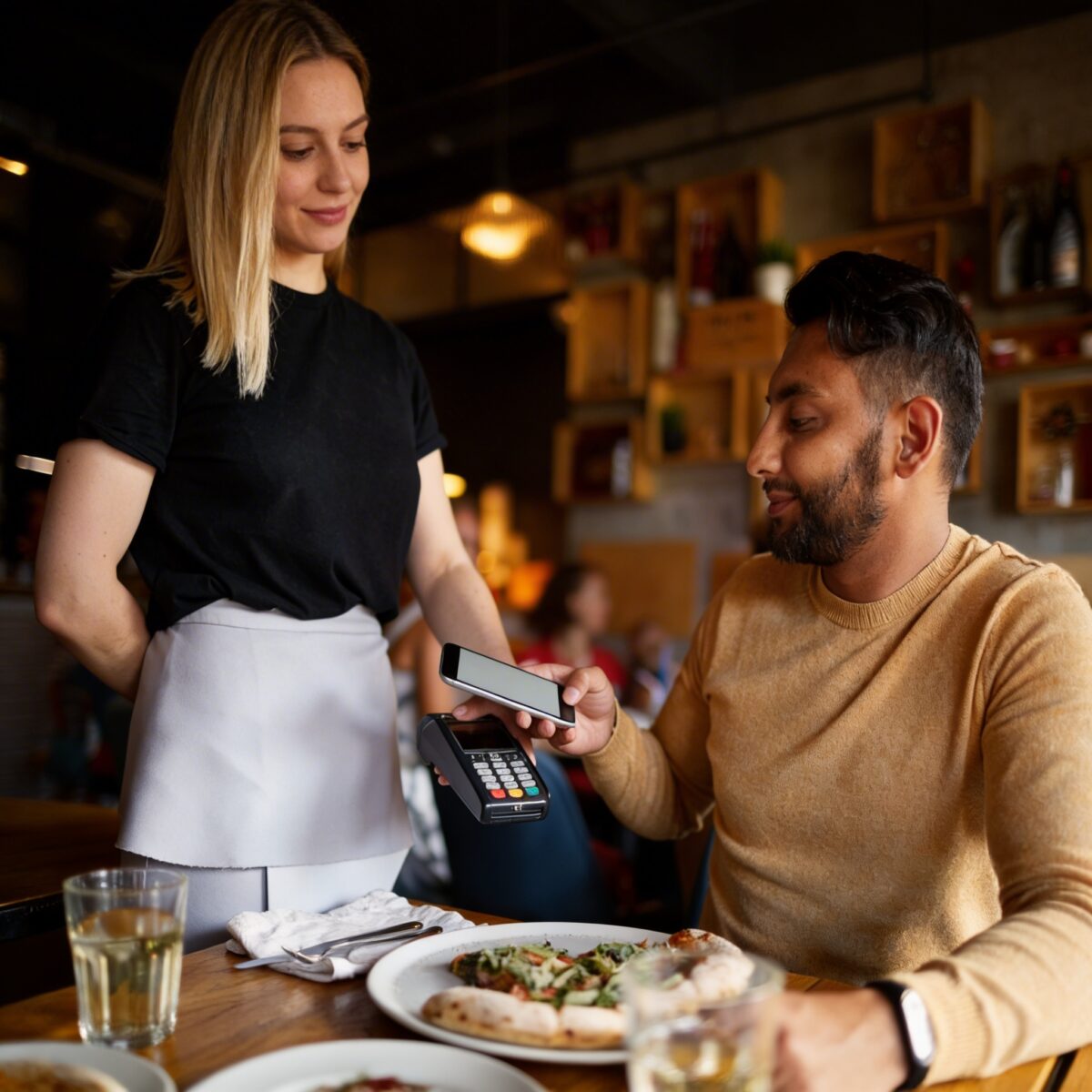 Customer making contactless payment with smartphone at restaurant table using card terminal